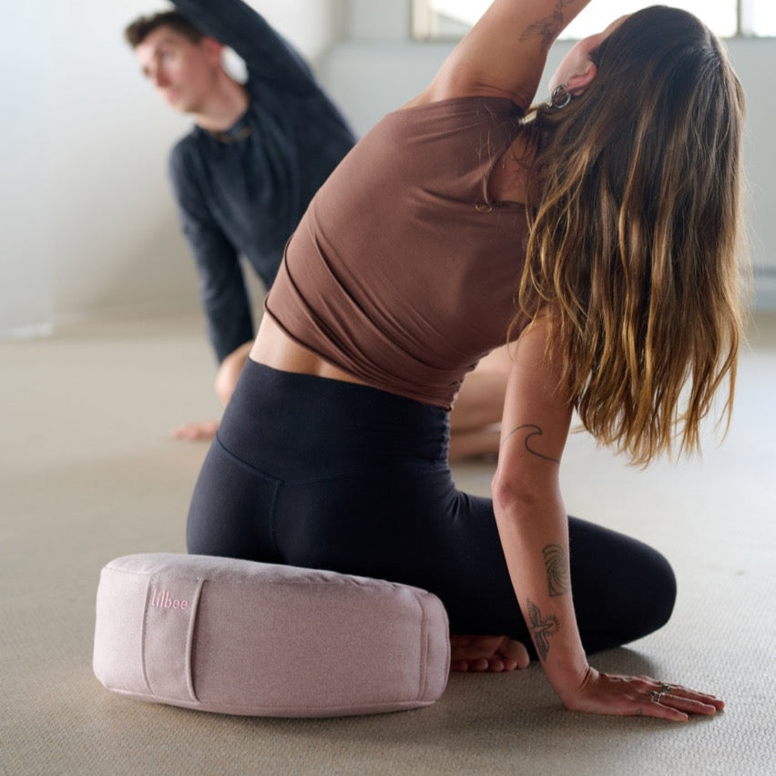 A woman performs a side stretch on a Lilbee pink zafu during a yoga class. Ergonomic meditation cushion supporting the spine and promoting balance, handmade in Canada.