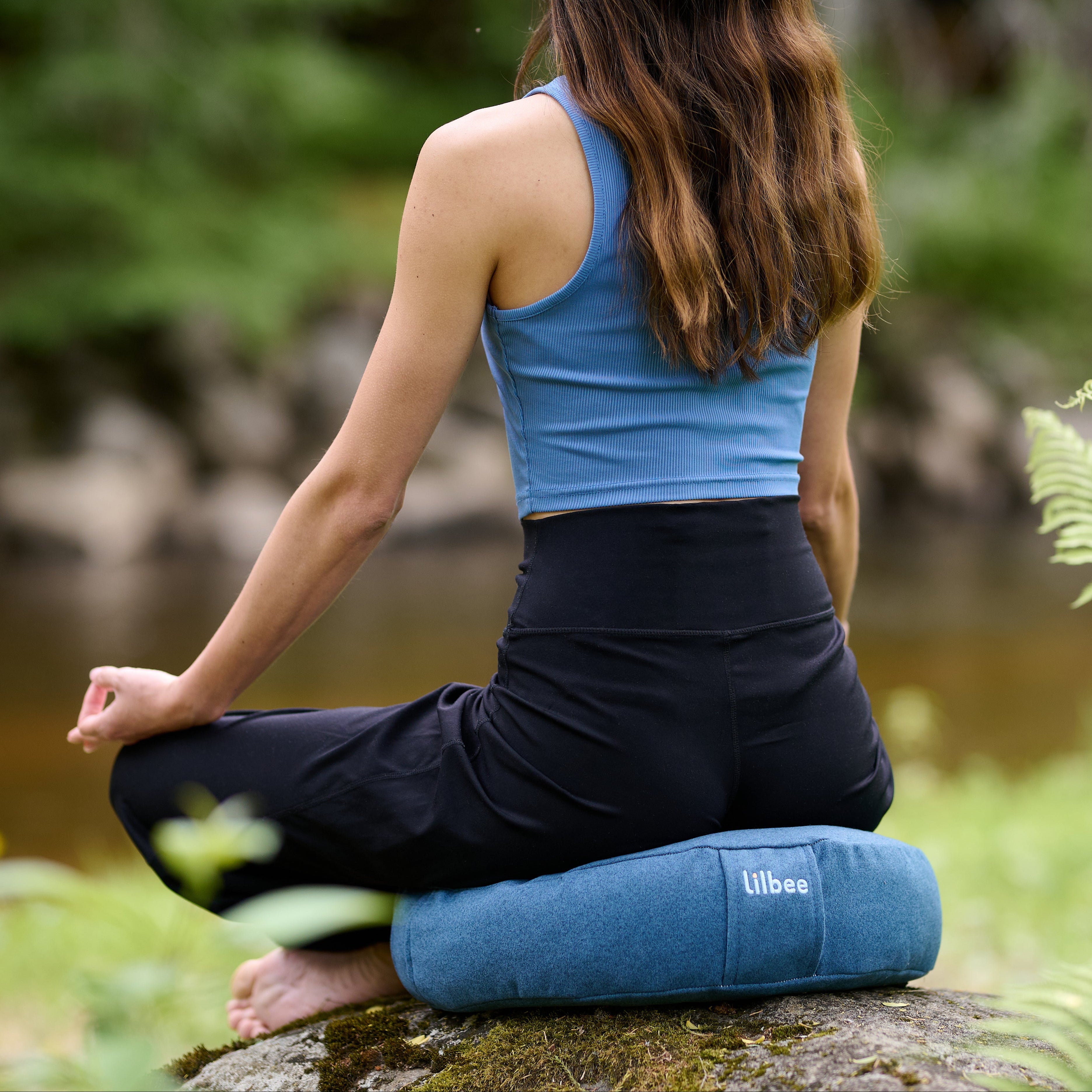 Woman meditating outdoors on a blue Lilbee Zafu cushion near water and green ferns.