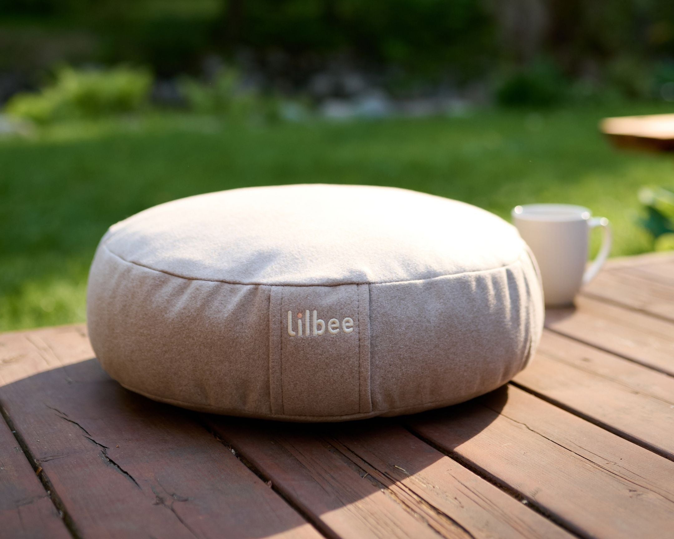 Beige Lilbee Low Zafu meditation cushion on a wooden outdoor table in a peaceful garden setting.