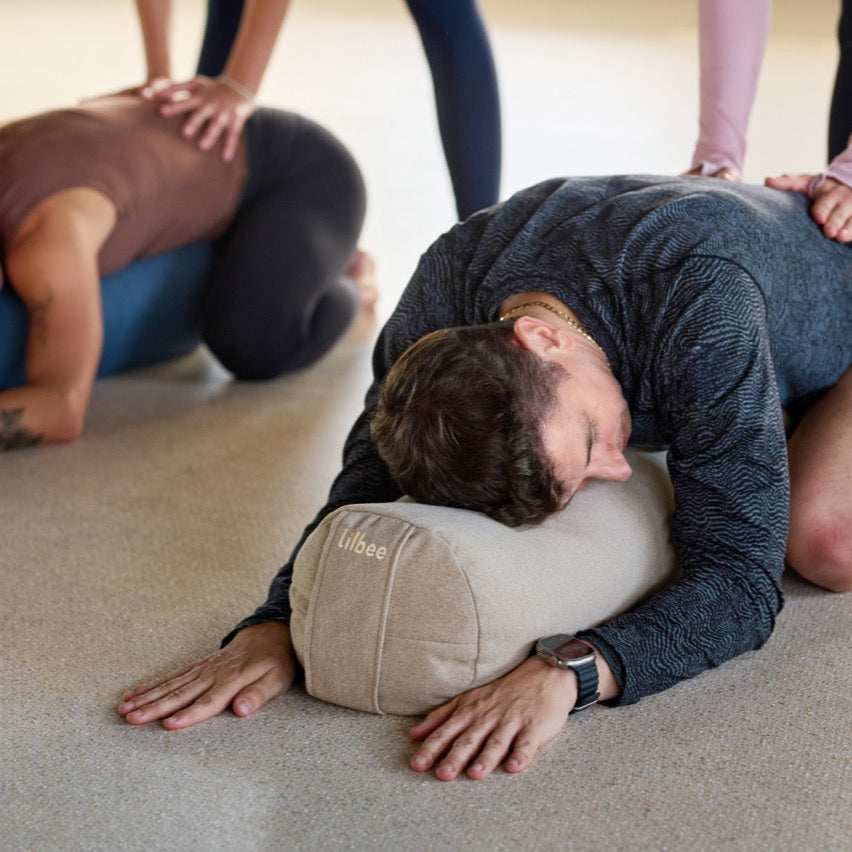 Adults resting forward on Lilbee round bolsters during a restorative yoga session. Cylindrical cushion providing balance and comfort, sustainably crafted in Canada with recycled filling.