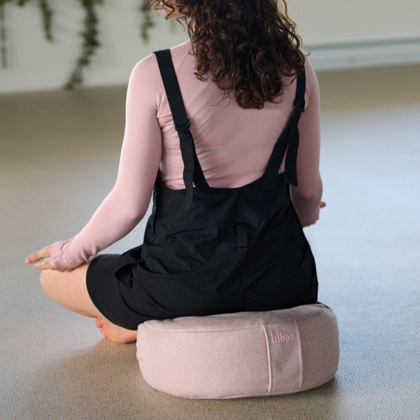Woman sitting peacefully on a blush pink Lilbee zafu in a bright yoga studio. Minimalist meditation cushion made in Canada from recycled fabric scraps for eco-conscious comfort.