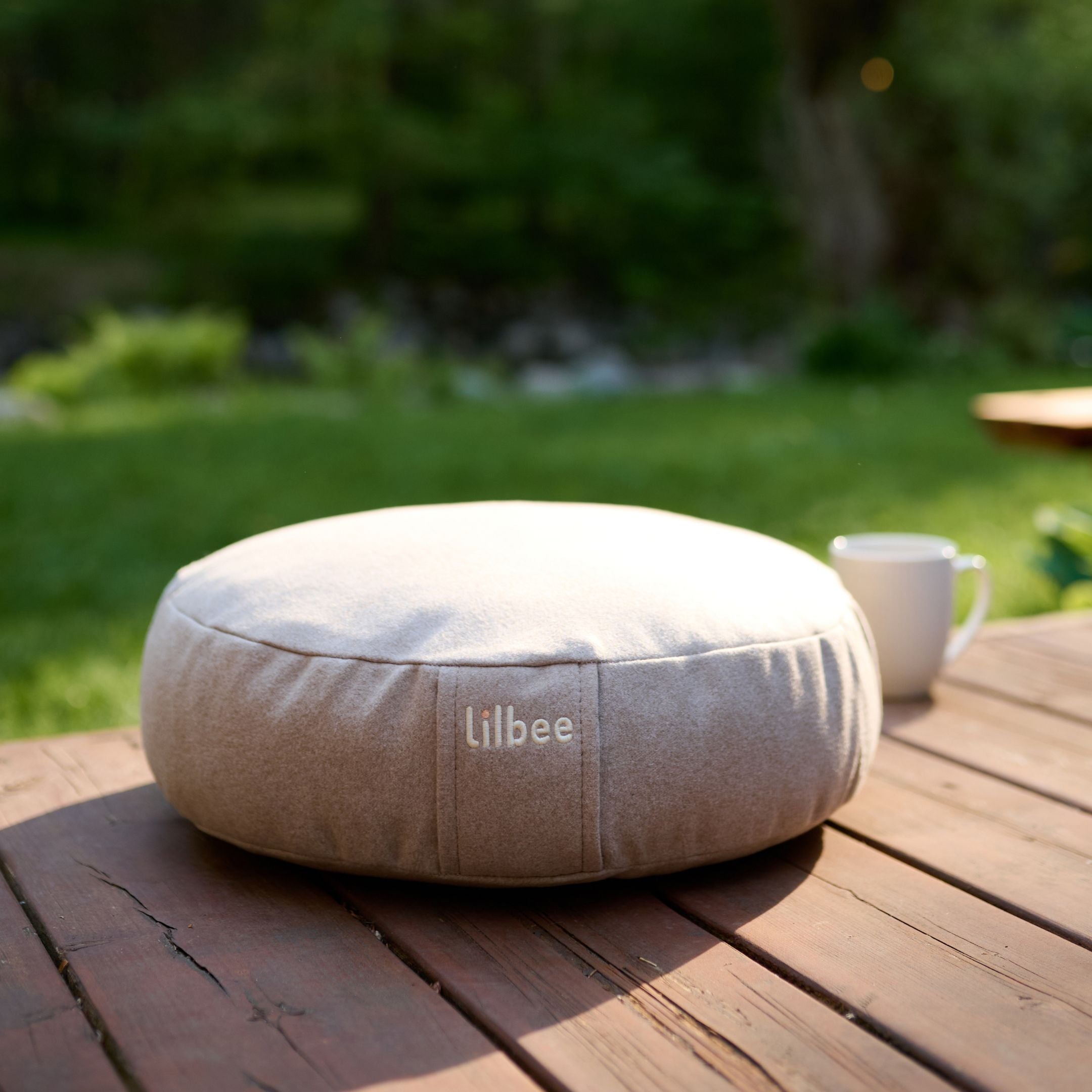 Beige Lilbee Low Zafu meditation cushion on a wooden outdoor table in a peaceful garden setting.